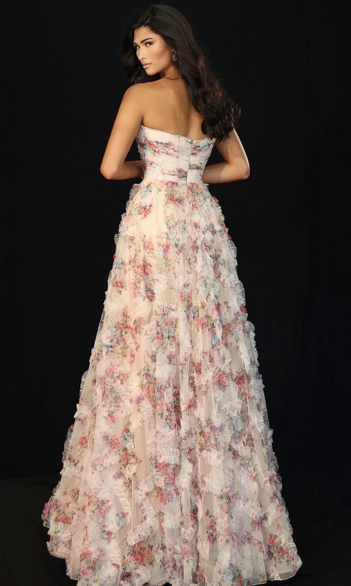 A woman wearing the Jovani 46783 long prom dress, featuring a strapless floral design and ruffle organza skirt, stands with her back to the camera against a black background.